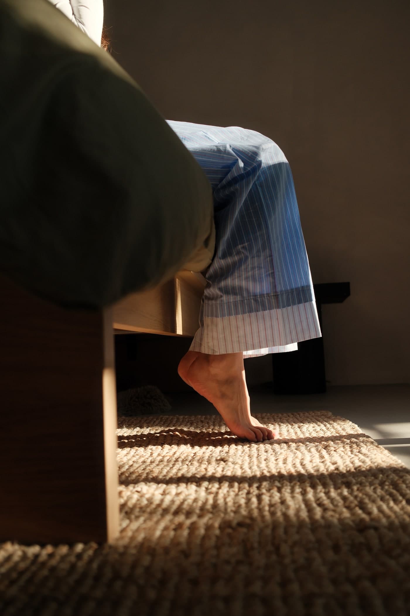 Woman stepping barefoot out of bed onto a woven rug, morning light